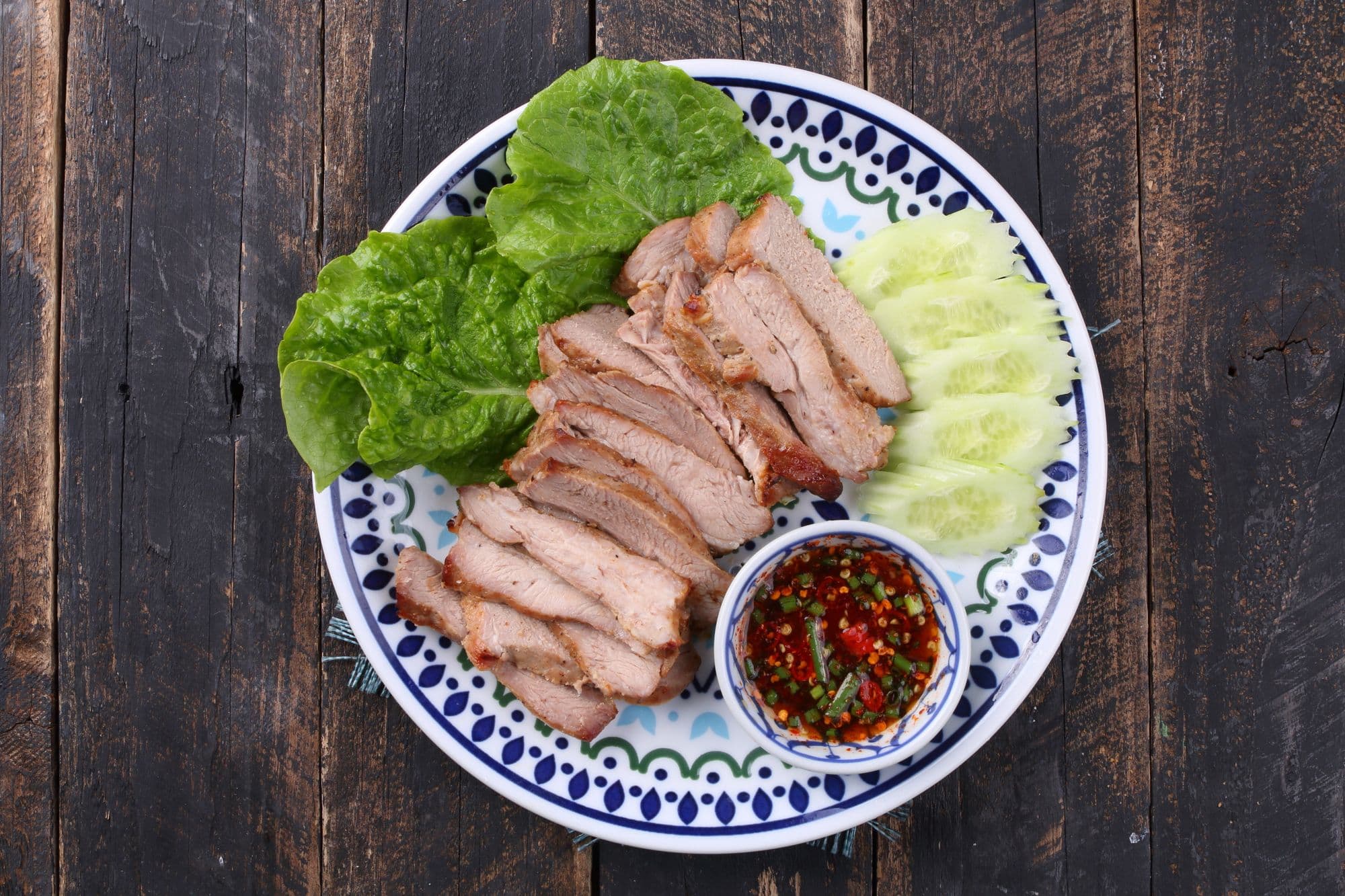 Sliced grilled pork on a blue-patterned Thai plate with lettuce, cucumber and a dish of chilli sauce, on dark wood