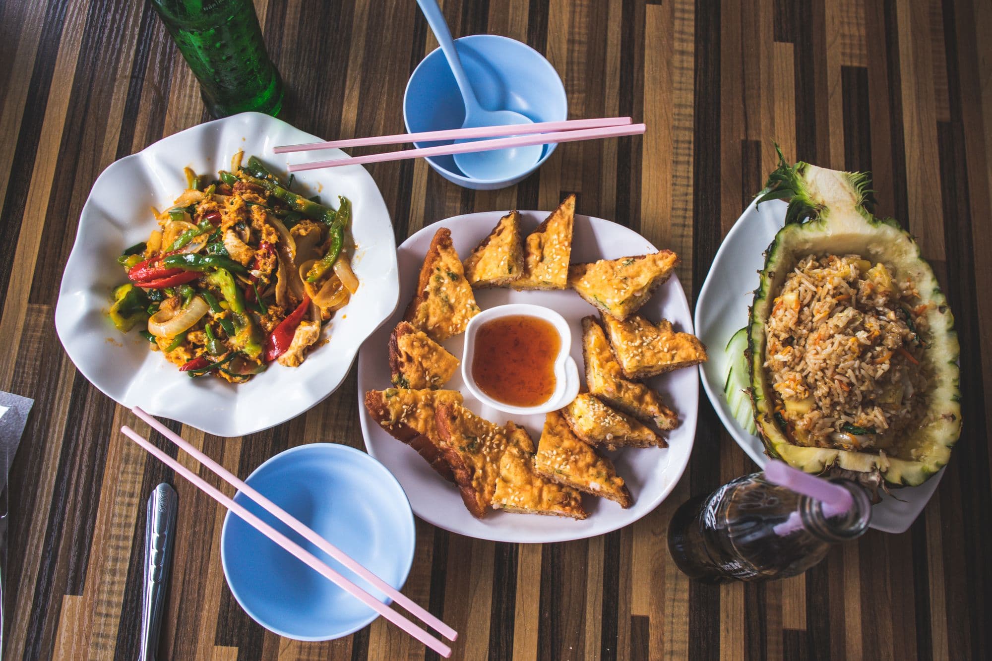 Several dishes photographed from directly above on a wooden table: a stir-fry, fried tofu and rice served in a pineapple