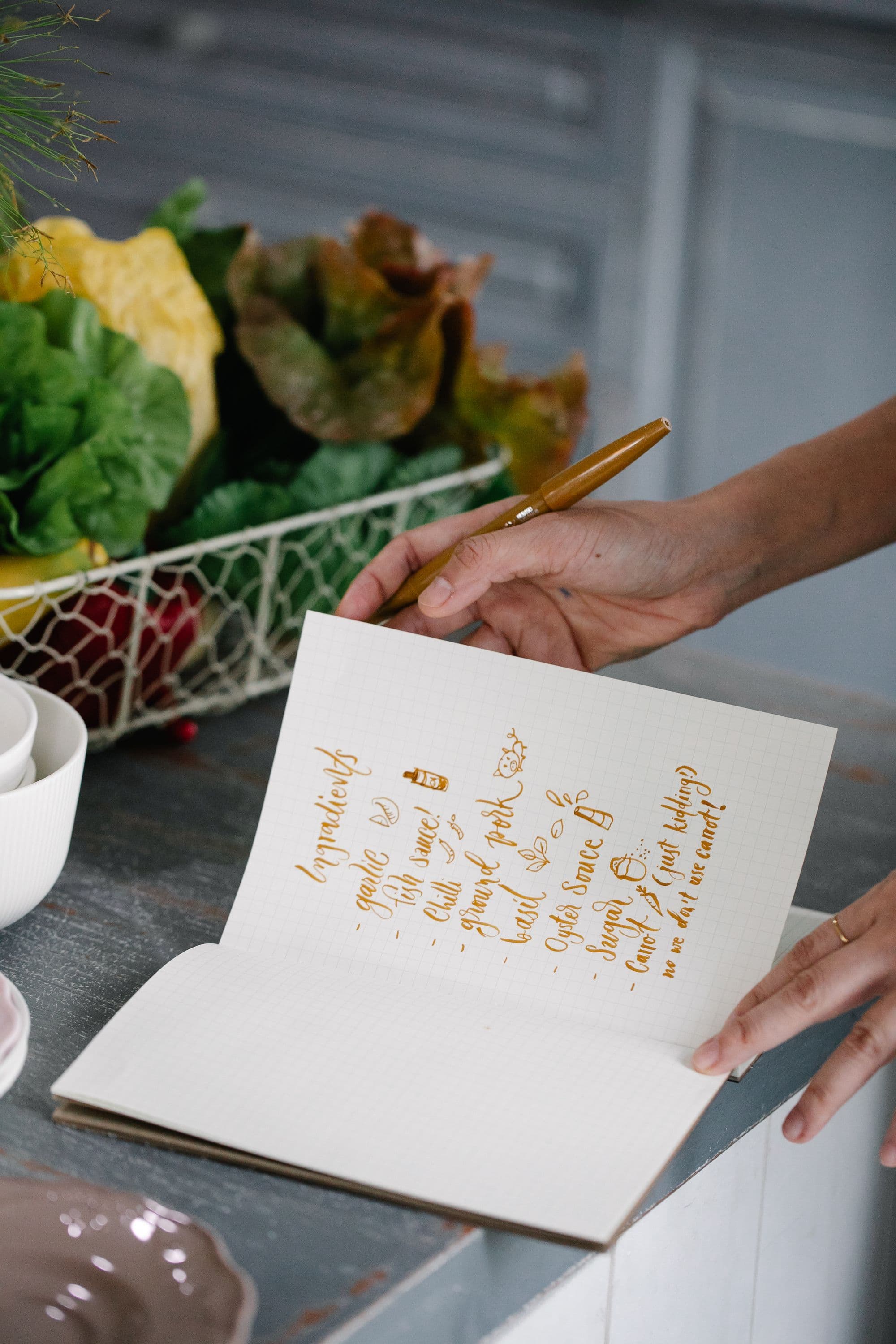 Two hands writing a list of ingredients in an open notebook beside garlic and chillies