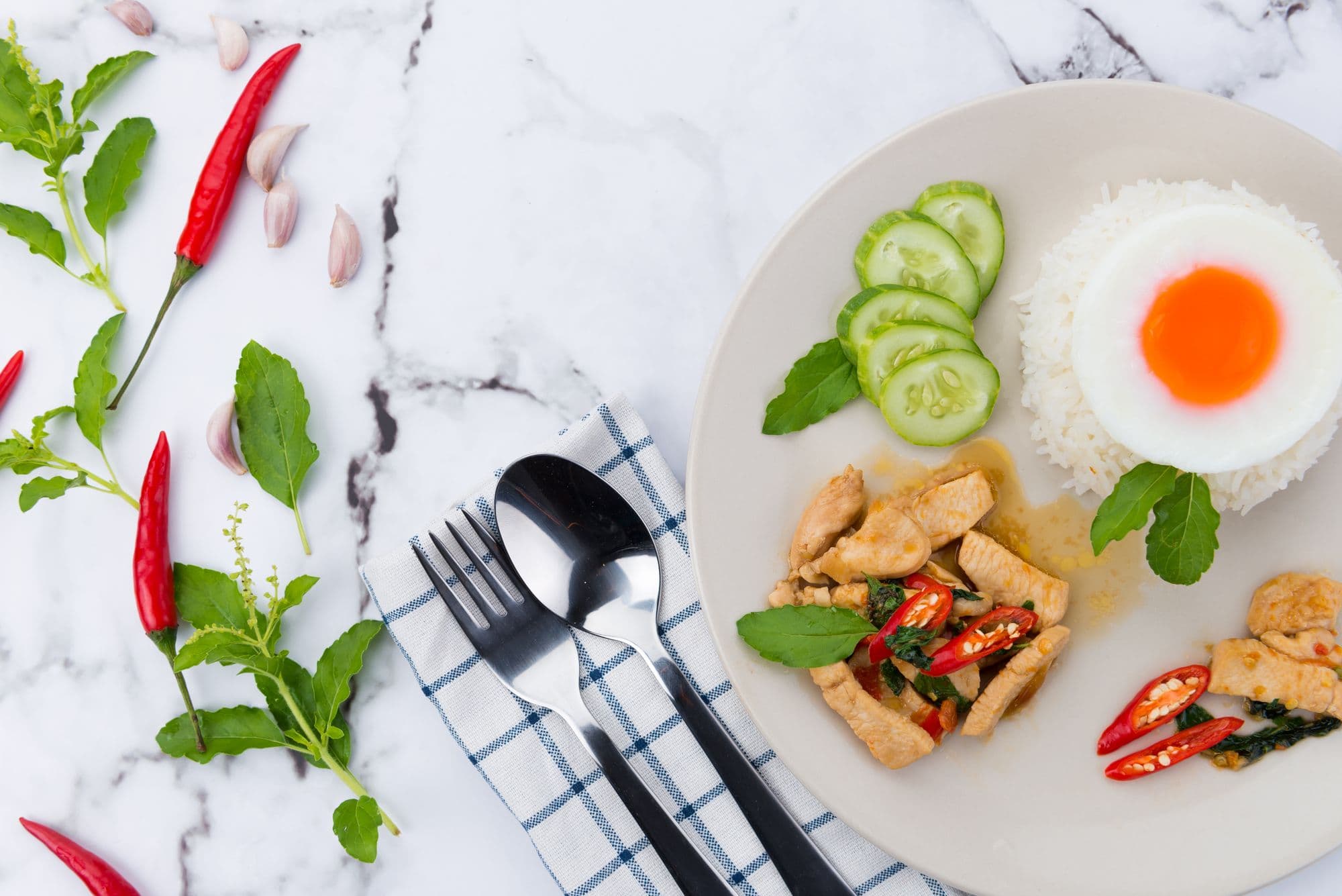 Thai basil chicken with jasmine rice and a fried egg on a marble bench, with fresh chillies, garlic and holy basil laid out beside it