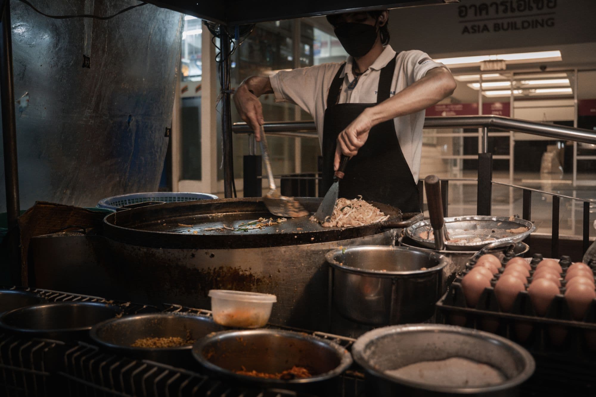 A cook working a wok over a burner in a busy kitchen, turned away from the camera