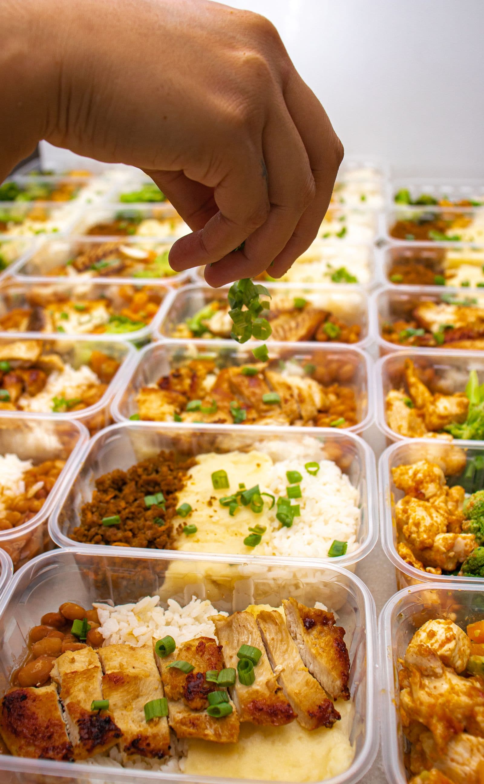 A cook scattering sliced spring onion over rows of meal-prep containers of chicken, rice and vegetables